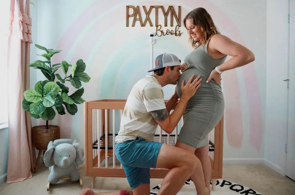 הורים לעתיד ברגע מרגש של ציפייה בחדר התינוקות, הכולל מיטת תינוק מעוצבת, פיל נדנדה ופריטי דקורציה משלימים מבית מוצצים.